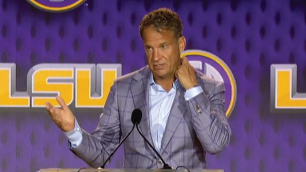 Man in a gray plaid suit speaks at a podium on a purple LSU-themed stage, gesturing with his left hand.