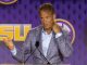 Man in a gray plaid suit speaks at a podium on a purple LSU-themed stage, gesturing with his left hand.