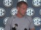 Man in a gray athletic shirt speaking at a press conference, standing behind a microphone with SEC logos on the backdrop