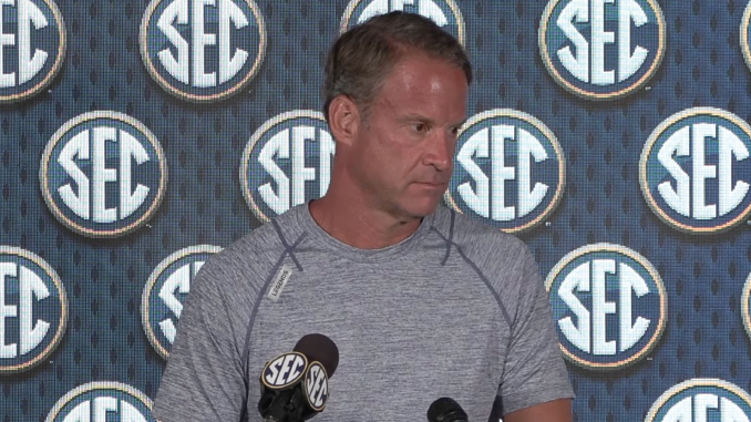 Man in a gray athletic shirt speaking at a press conference, standing behind a microphone with SEC logos on the backdrop