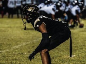 American football player in a black uniform crouching in a ready stance on a grassy field during a game.