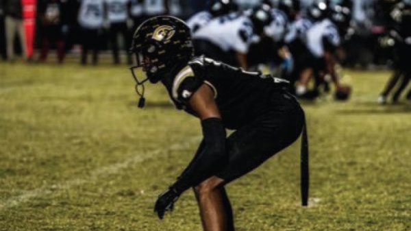 American football player in a black uniform crouching in a ready stance on a grassy field during a game.