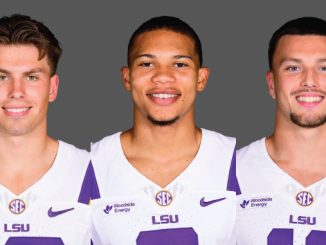 Three LSU football players in white and purple jerseys stand side by side, smiling at the camera against a gray backdrop.