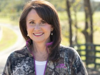 Smiling woman with dark hair and pearl earrings, wearing a camo jacket on a sunny rural path with a fence in the background.