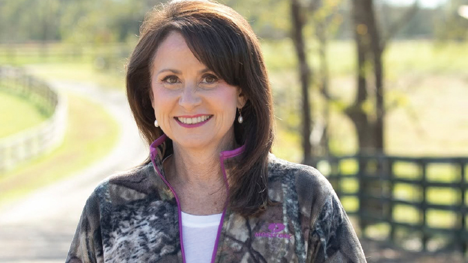 Smiling woman with dark hair and pearl earrings, wearing a camo jacket on a sunny rural path with a fence in the background.