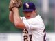 Baseball pitcher in a white uniform and dark cap, glove raised as he prepares to throw.