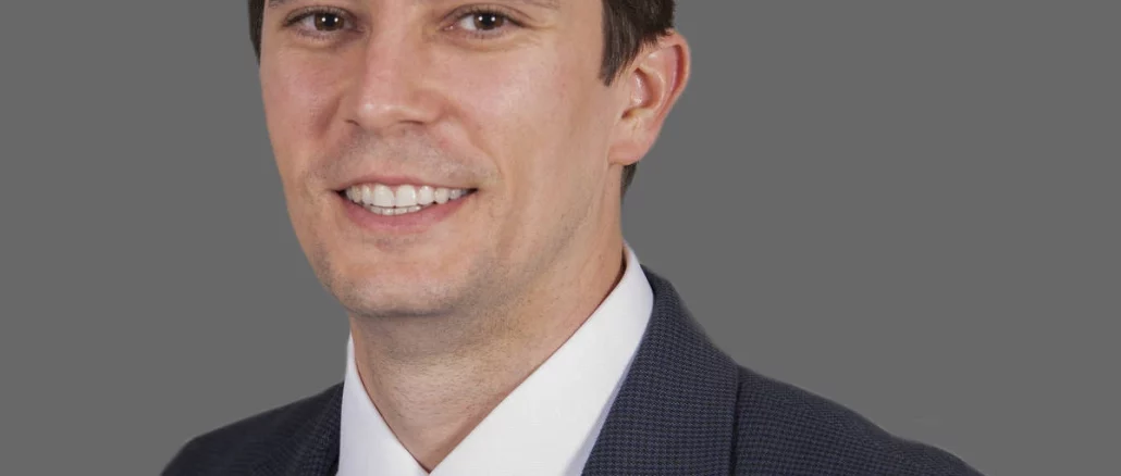 Professional head-and-shoulders portrait of a smiling man in a dark suit and light blue tie against a gray background.