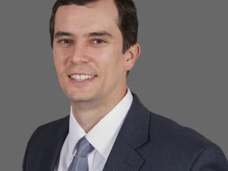 Professional head-and-shoulders portrait of a smiling man in a dark suit and light blue tie against a gray background.