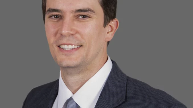 Professional head-and-shoulders portrait of a smiling man in a dark suit and light blue tie against a gray background.