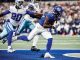 New York Giants running back in blue jersey sprinting with the ball as a Cowboys defender chases him on the field.