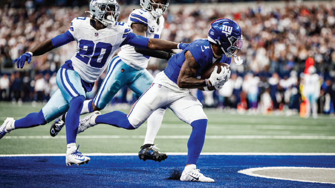 New York Giants running back in blue jersey sprinting with the ball as a Cowboys defender chases him on the field.
