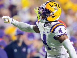 LSU football player wearing a white jersey with purple numbers and a yellow helmet (number 4) points during a game on the field.