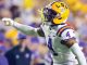 LSU football player wearing a white jersey with purple numbers and a yellow helmet (number 4) points during a game on the field.