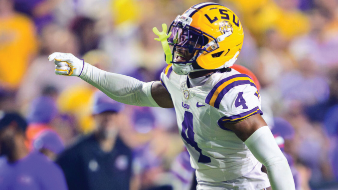 LSU football player wearing a white jersey with purple numbers and a yellow helmet (number 4) points during a game on the field.