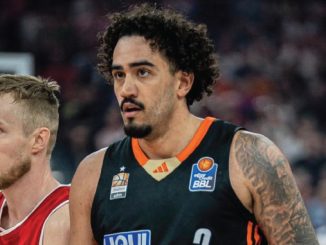 Two basketball players on the court during a game; foreground player wears a black jersey with orange trim, behind him a red-jersey opponent stands close by.