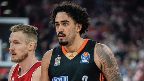 Two basketball players on the court during a game; foreground player wears a black jersey with orange trim, behind him a red-jersey opponent stands close by.