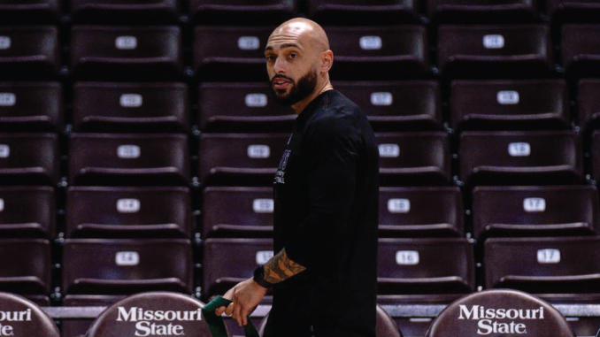 MarcusBrock_MissouriState | TigerRag.com A bald, bearded man in a black shirt stands in an arena concourse with empty maroon seats behind him, near a Missouri State bench area.
