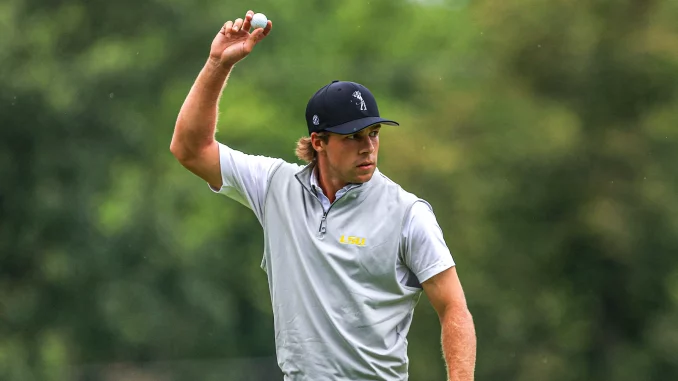 Male golfer in a light gray LSU polo raises his right arm, tossing a golf ball on a green course.