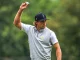 Male golfer in a light gray LSU polo raises his right arm, tossing a golf ball on a green course.