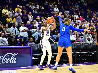 LSU player in white #23 drives with the ball while defender in blue #0 reaches in during a college basketball game with a packed crowd behind.