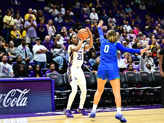 LSU player in white #23 drives with the ball while defender in blue #0 reaches in during a college basketball game with a packed crowd behind.