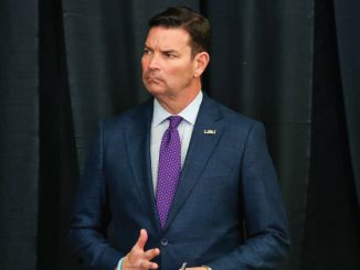 Man in a navy suit with a purple tie stands on a stage with black curtains, mid-gesture and serious expression, wearing an LSU pin on his lapel.