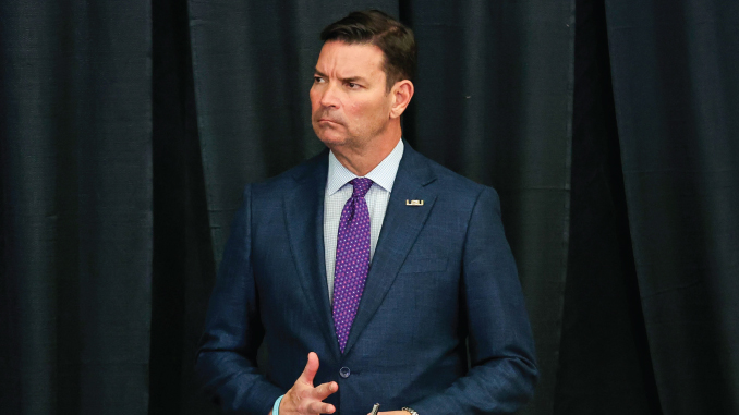 Man in a navy suit with a purple tie stands on a stage with black curtains, mid-gesture and serious expression, wearing an LSU pin on his lapel.