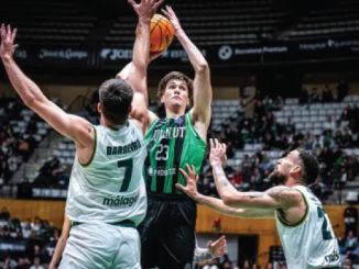 Basketball player in a green jersey jumps to shoot, surrounded by two white-jersey defenders in a crowded arena.