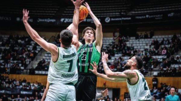 Basketball player in a green jersey jumps to shoot, surrounded by two white-jersey defenders in a crowded arena.