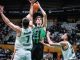 Basketball player in a green jersey jumps to shoot, surrounded by two white-jersey defenders in a crowded arena.