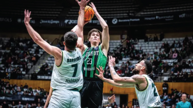 Basketball player in a green jersey jumps to shoot, surrounded by two white-jersey defenders in a crowded arena.