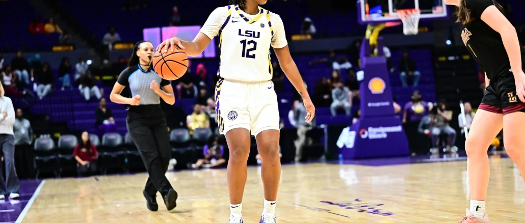 LSU women’s basketball player wearing #12 in a white jersey dribbles the ball while a defender in black guards her near the court with a referee and spectators in the background.