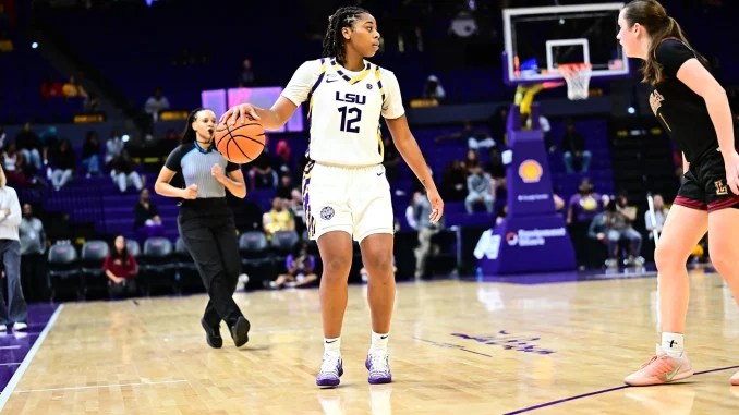 LSU women’s basketball player wearing #12 in a white jersey dribbles the ball while a defender in black guards her near the court with a referee and spectators in the background.