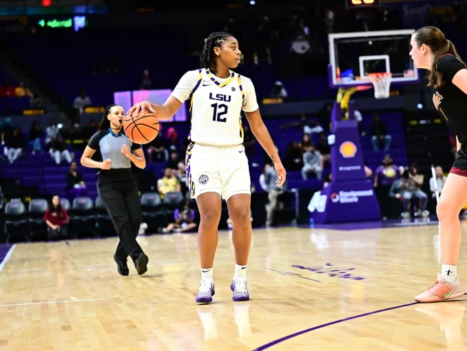 LSU women’s basketball player wearing #12 in a white jersey dribbles the ball while a defender in black guards her near the court with a referee and spectators in the background.