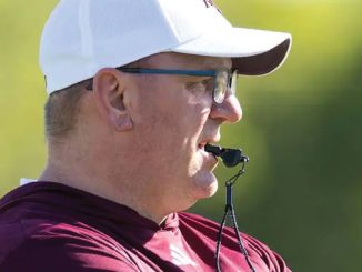 Profile view of a man wearing a white cap and glasses, with a whistle in his mouth, outdoors in a maroon shirt.