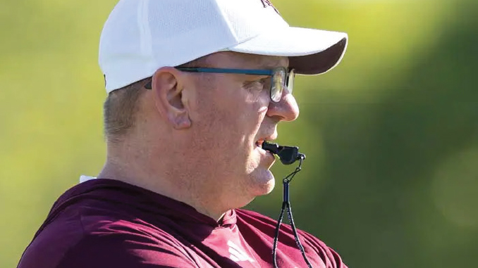 Profile view of a man wearing a white cap and glasses, with a whistle in his mouth, outdoors in a maroon shirt.