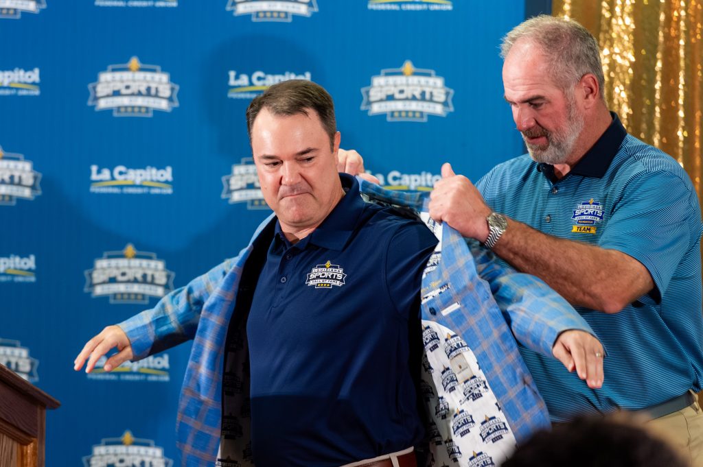 Two men at a ceremony wearing matching team shirts; one helps the other put on a blue plaid jacket, backdrop branded with sports logos.