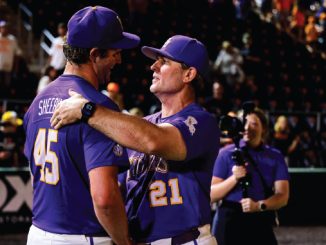 Two baseball players in purple uniforms hug on the field as fans watch in the stands, numbers 45 and 21 visible on their jerseys.