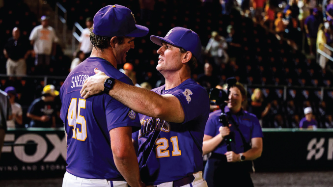 Two baseball players in purple uniforms hug on the field as fans watch in the stands, numbers 45 and 21 visible on their jerseys.