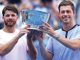 Two male tennis players hold a large silver trophy together and smile at the camera on a blue court.