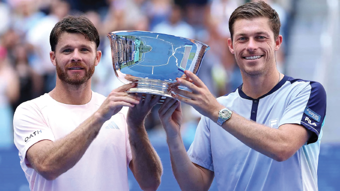 Two male tennis players hold a large silver trophy together and smile at the camera on a blue court.