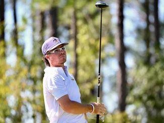 Golfer in a white polo and cap swings a driver on a sunlit forest course with trees in the background.