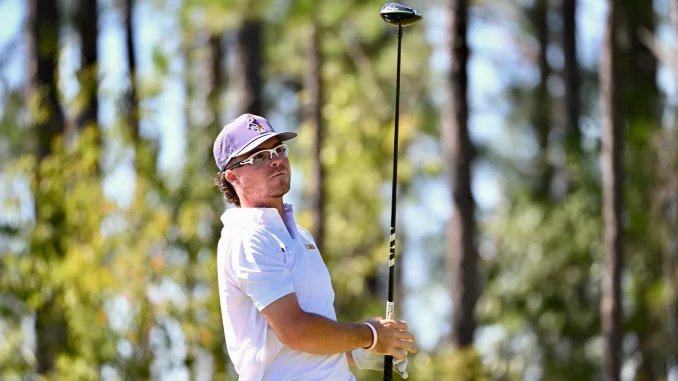 Golfer in a white polo and cap swings a driver on a sunlit forest course with trees in the background.