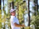 Golfer in a white polo and cap swings a driver on a sunlit forest course with trees in the background.