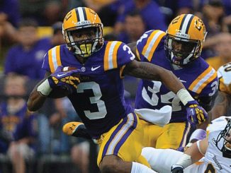 LSU Tigers football players in purple and gold run with the ball during a game, pursued by defenders in white uniforms.