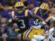 LSU Tigers football players in purple and gold run with the ball during a game, pursued by defenders in white uniforms.