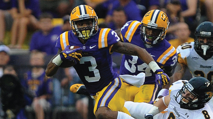 LSU Tigers football players in purple and gold run with the ball during a game, pursued by defenders in white uniforms.