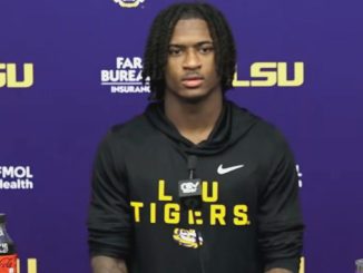 Young man with braided hair in a black LSU Tigers hoodie at a press conference, purple backdrop with LSU logos.