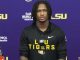 Young man with braided hair in a black LSU Tigers hoodie at a press conference, purple backdrop with LSU logos.