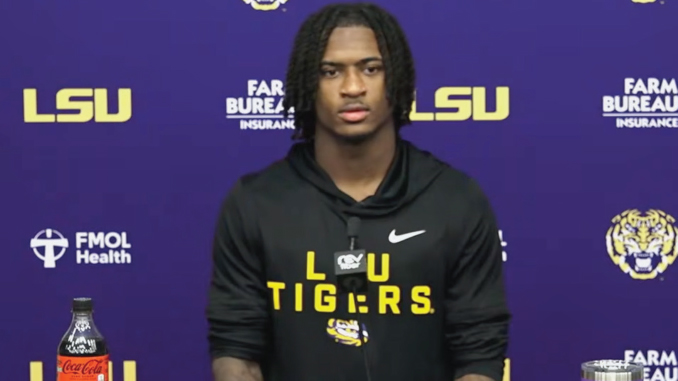 Young man with braided hair in a black LSU Tigers hoodie at a press conference, purple backdrop with LSU logos.